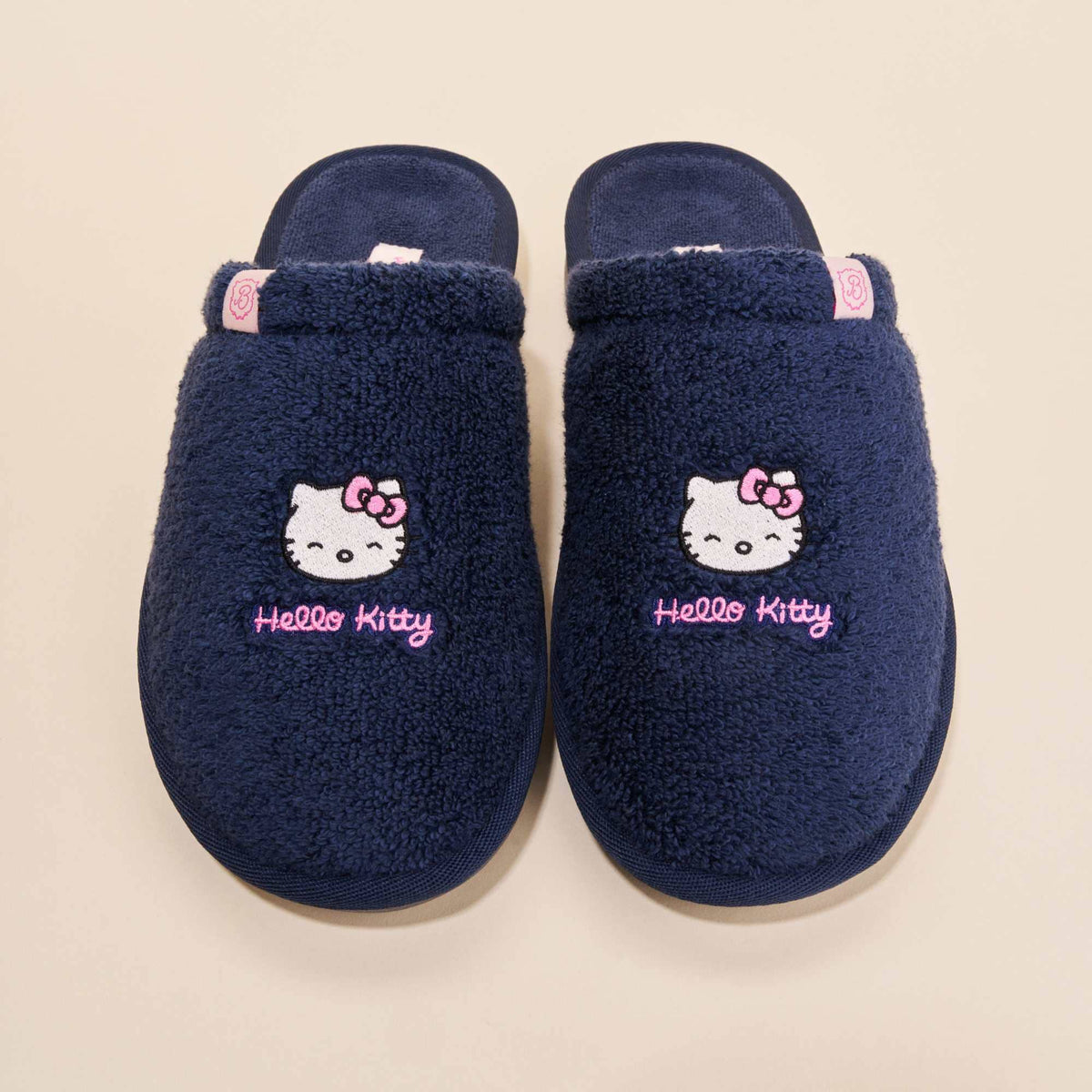 Top-down view of navy terry Hello Kitty slippers with embroidered faces and pink Hello Kitty script on beige background