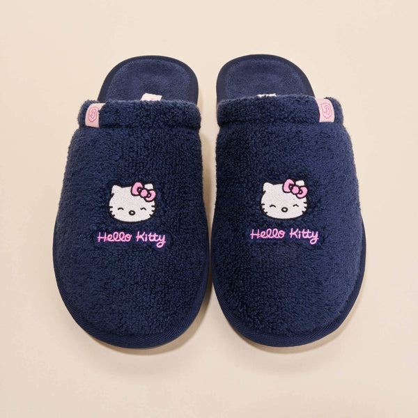 Top-down view of navy terry Hello Kitty slippers with embroidered faces and pink Hello Kitty script on beige background