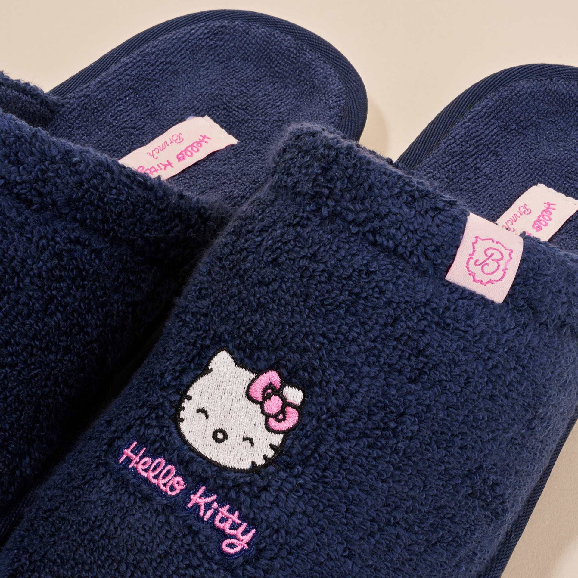 Top close-up of navy Hello Kitty terry slippers showing embroidered Hello Kitty face and pink 'Hello Kitty' script on beige background