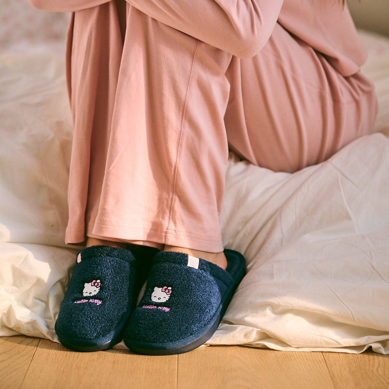 Front view of navy terry slip-on slippers with embroidered Hello Kitty, worn by a person sitting on a bed