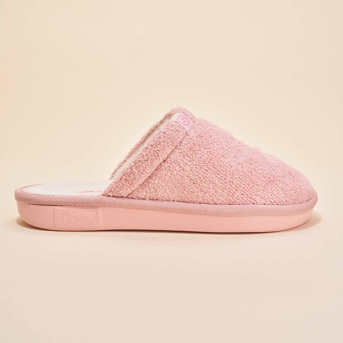 Side view of pink Hello Kitty terry slipper showing plush terry upper, embroidered Hello Kitty tag, and molded Brunch sole on a beige background