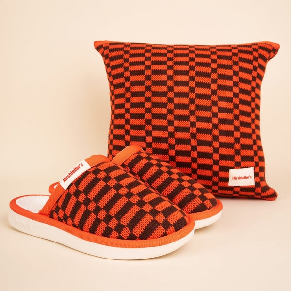 Three-quarter view of Amber Hirshleifers knit slippers showing orange and black checker knit upper and white foam sole on beige background