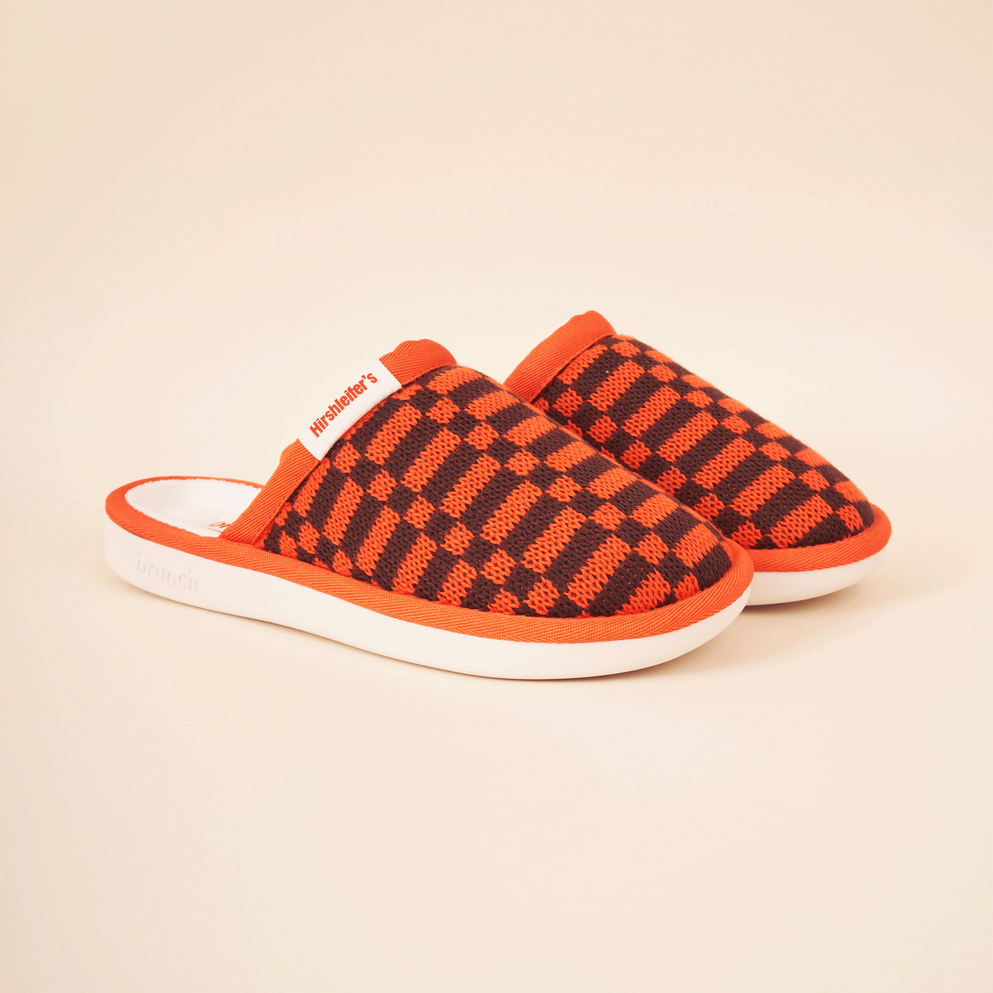 Three-quarter view of Amber knit checkered slip-on mule slipper showing orange-and-black knit upper, white cushioned sole, beige background