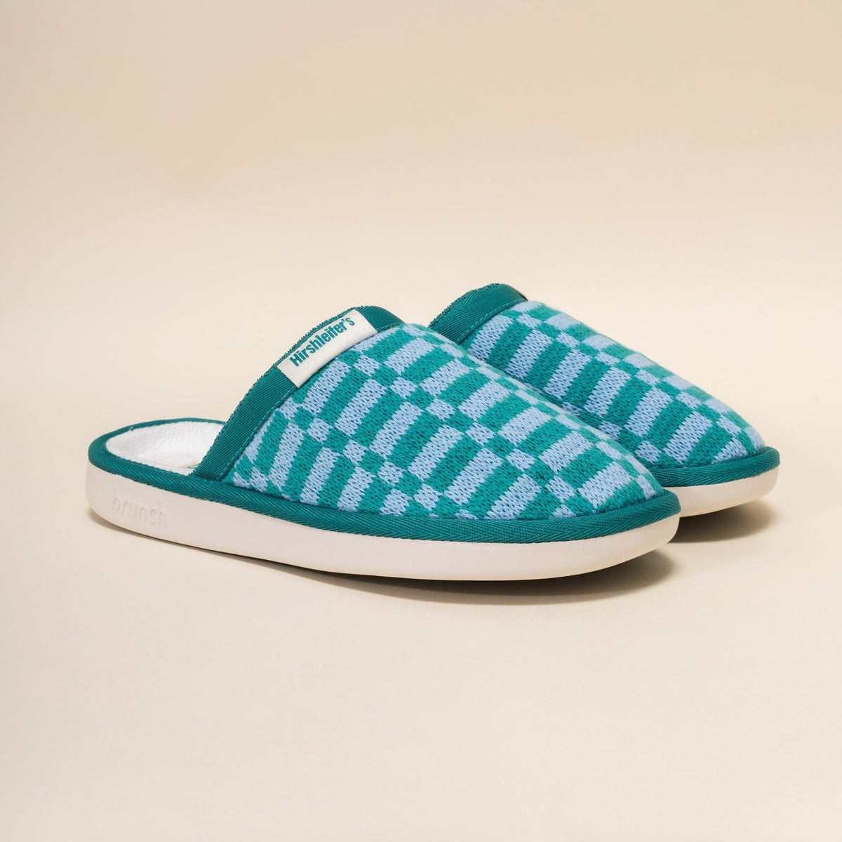 Three-quarter view of knit Hirshleifers slippers in marine green check with cushioned white sole and branded tag on beige