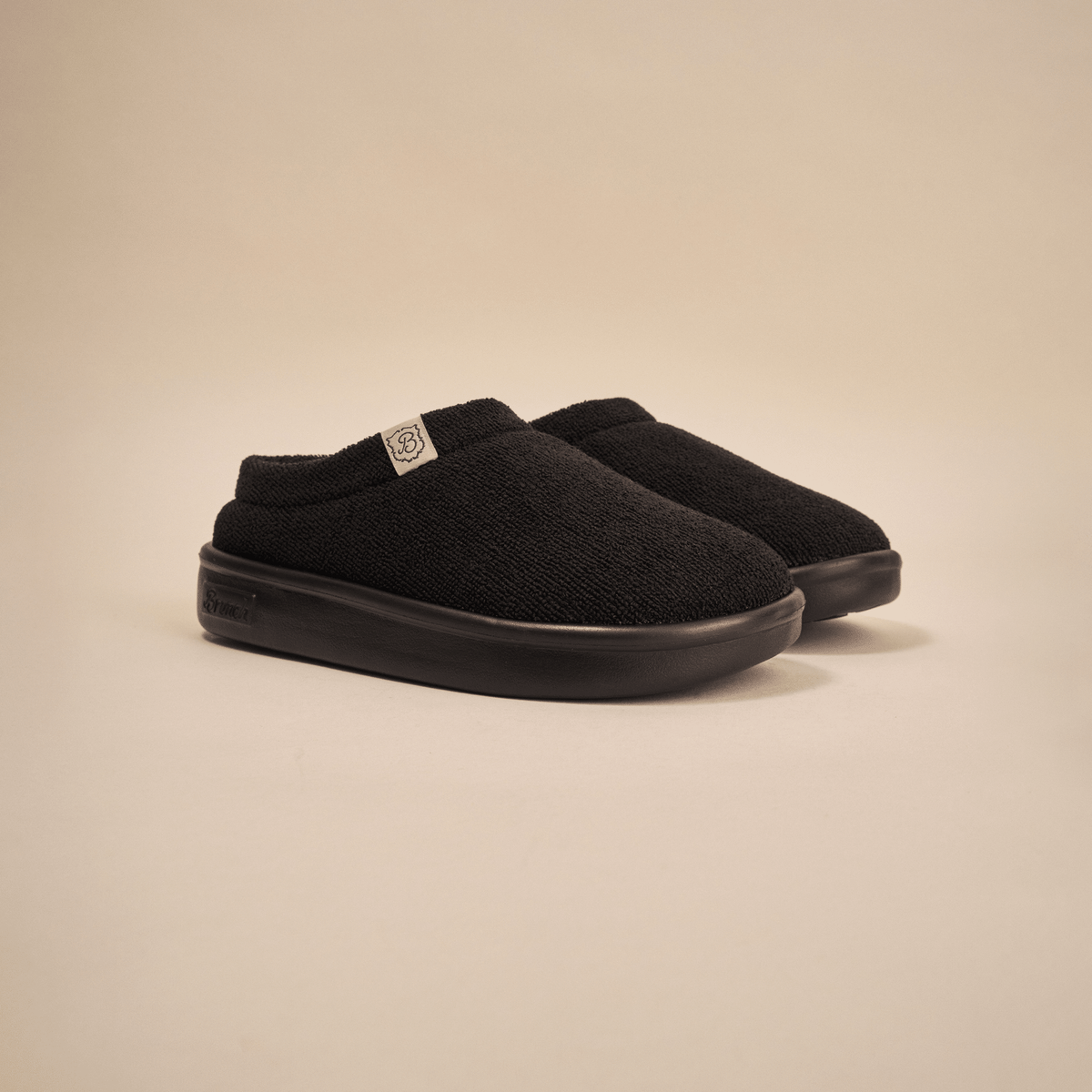 Front three-quarter view of Essential Kids slipper in black terry showing terry upper and EVA outsole on beige backdrop