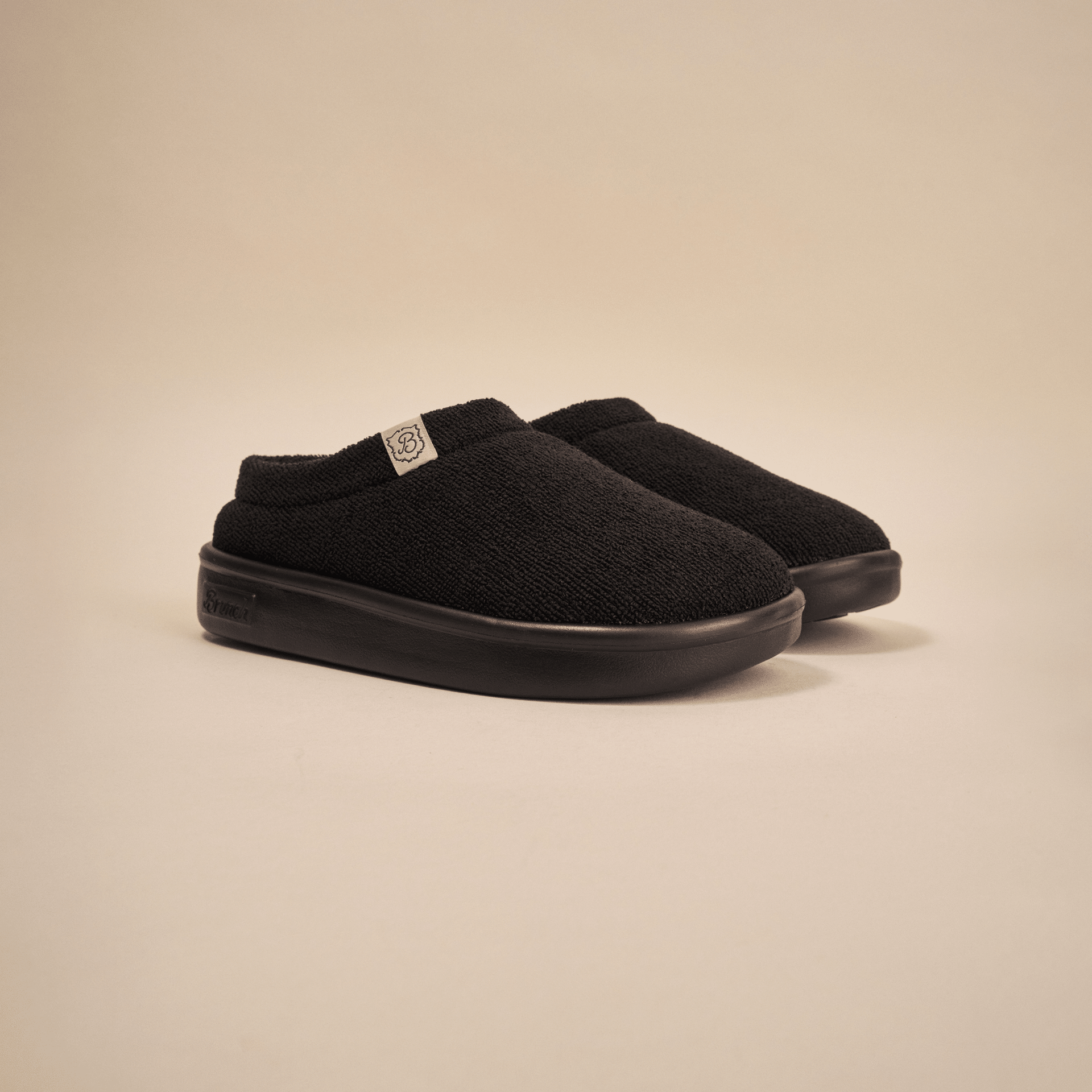 Front three-quarter view of Essential Kids slipper in black terry showing terry upper and EVA outsole on beige backdrop