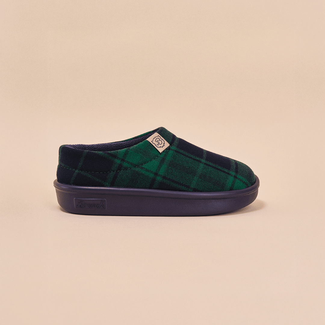 Side view of Essential Kids slipper in navy and green plaid showcasing slip-on silhouette and navy EVA outsole on light beige background