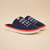 Front three-quarter view of navy Essential slipper featuring white embroidered "AMBER WAVES FARM AMAGANSETT", red trim and white sole on a beige background