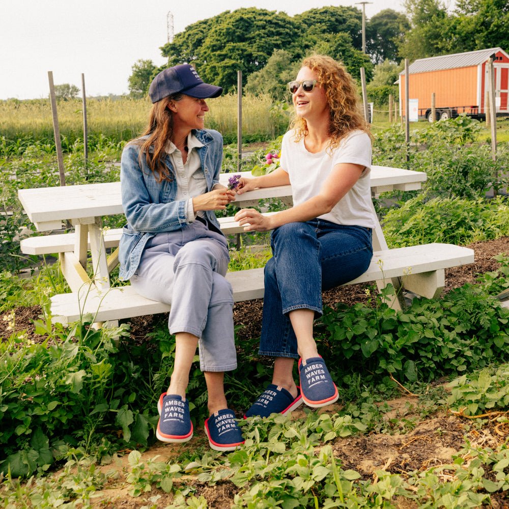 Two women sitting on a picnic bench at a farm, smiling and wearing navy Amber Waves Farm embroidered slippers