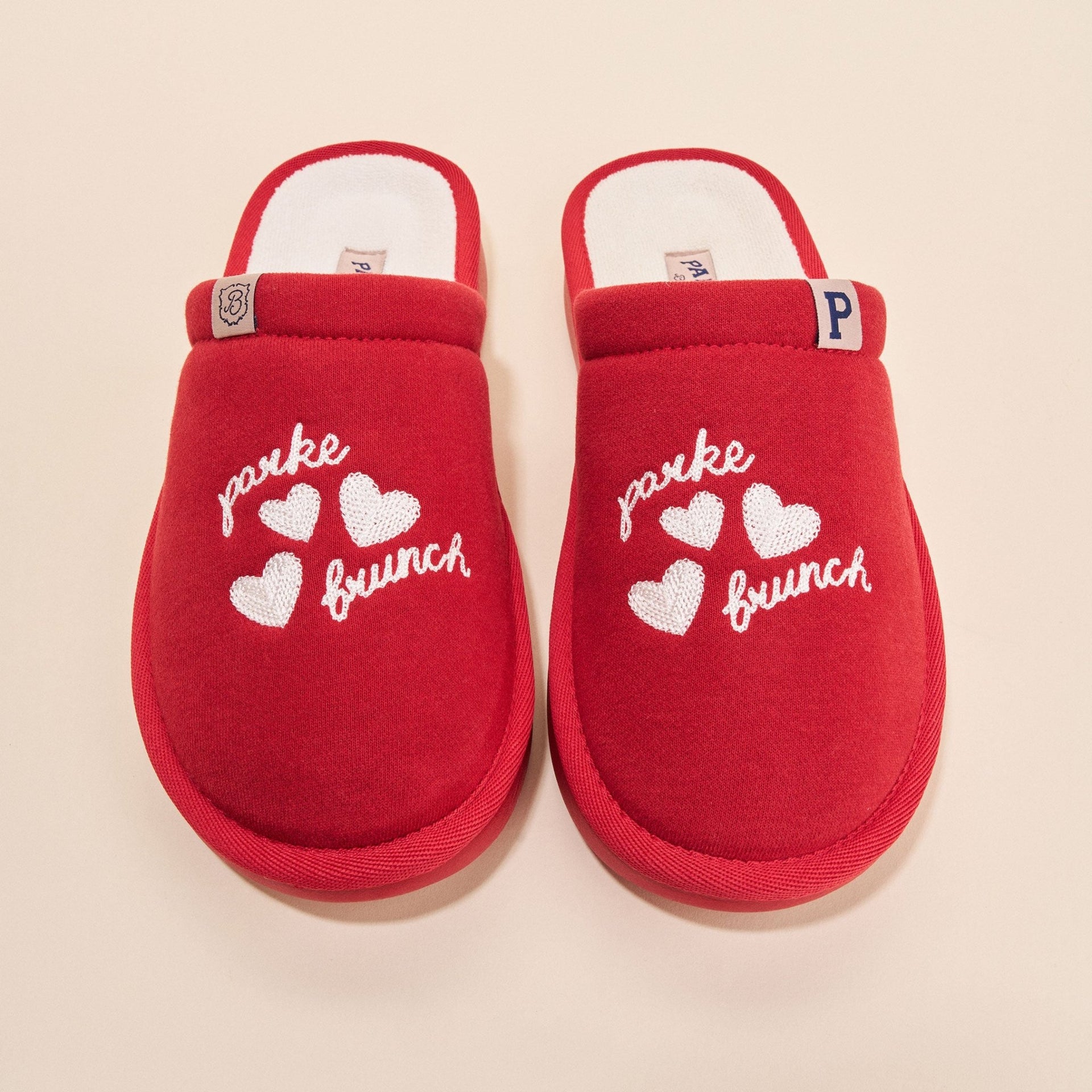 Top view of crimson Essential Parke fleece slippers showing white embroidered parke and brunch text, three hearts, and white lining on beige background