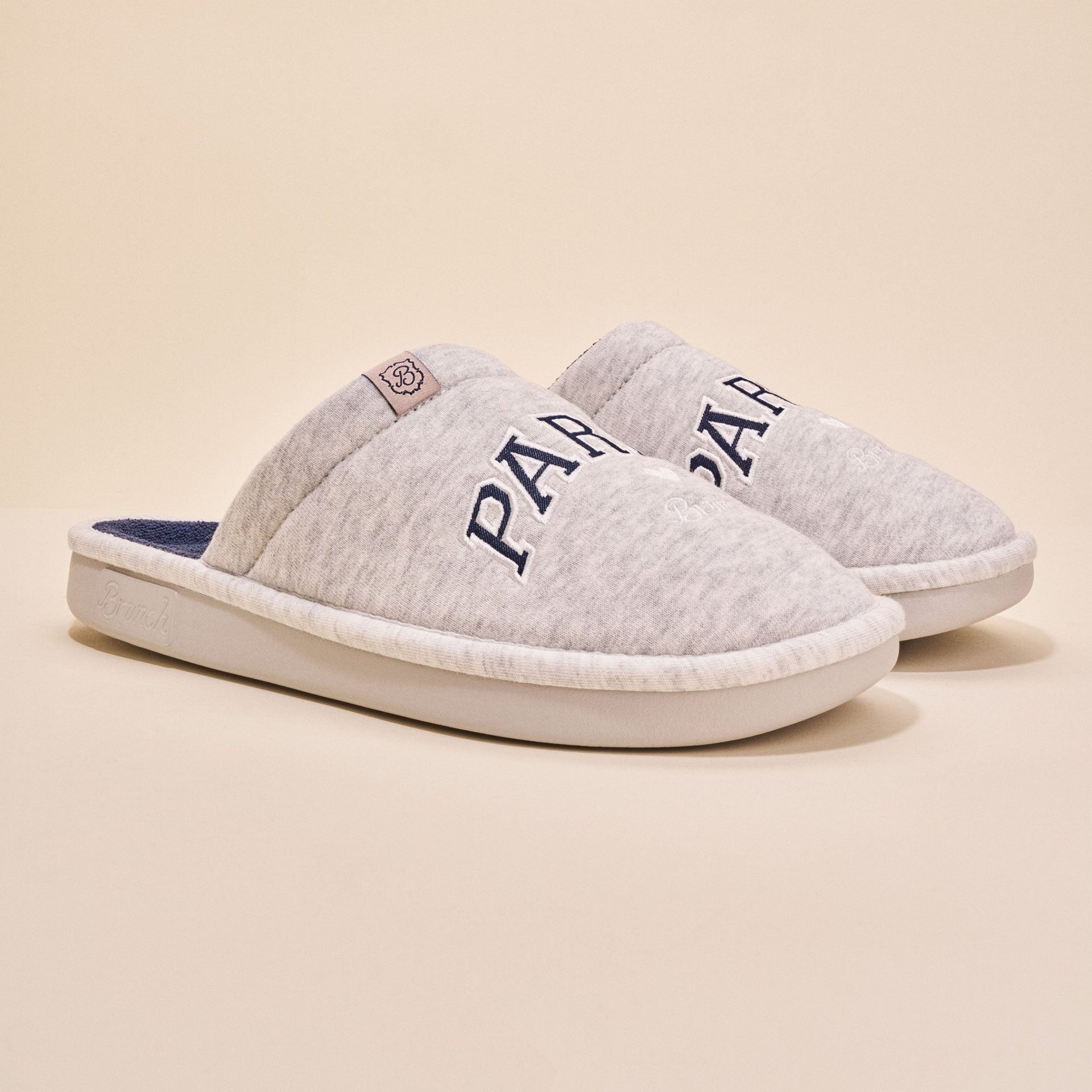 Angled view of pair of Essential Parke Varsity fleece slippers with embroidered PARKE lettering, cushioned white sole on beige
