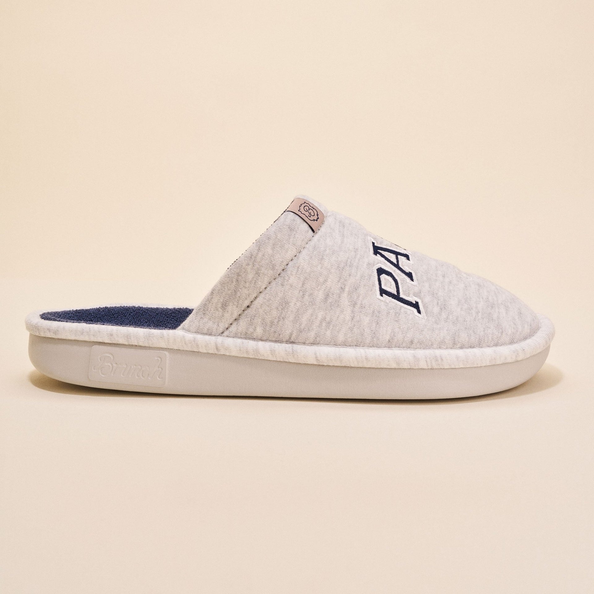 Side view of Essential Parke Varsity fleece slipper showcasing light gray fleece upper, navy terry lining, embroidered PA logo on beige background