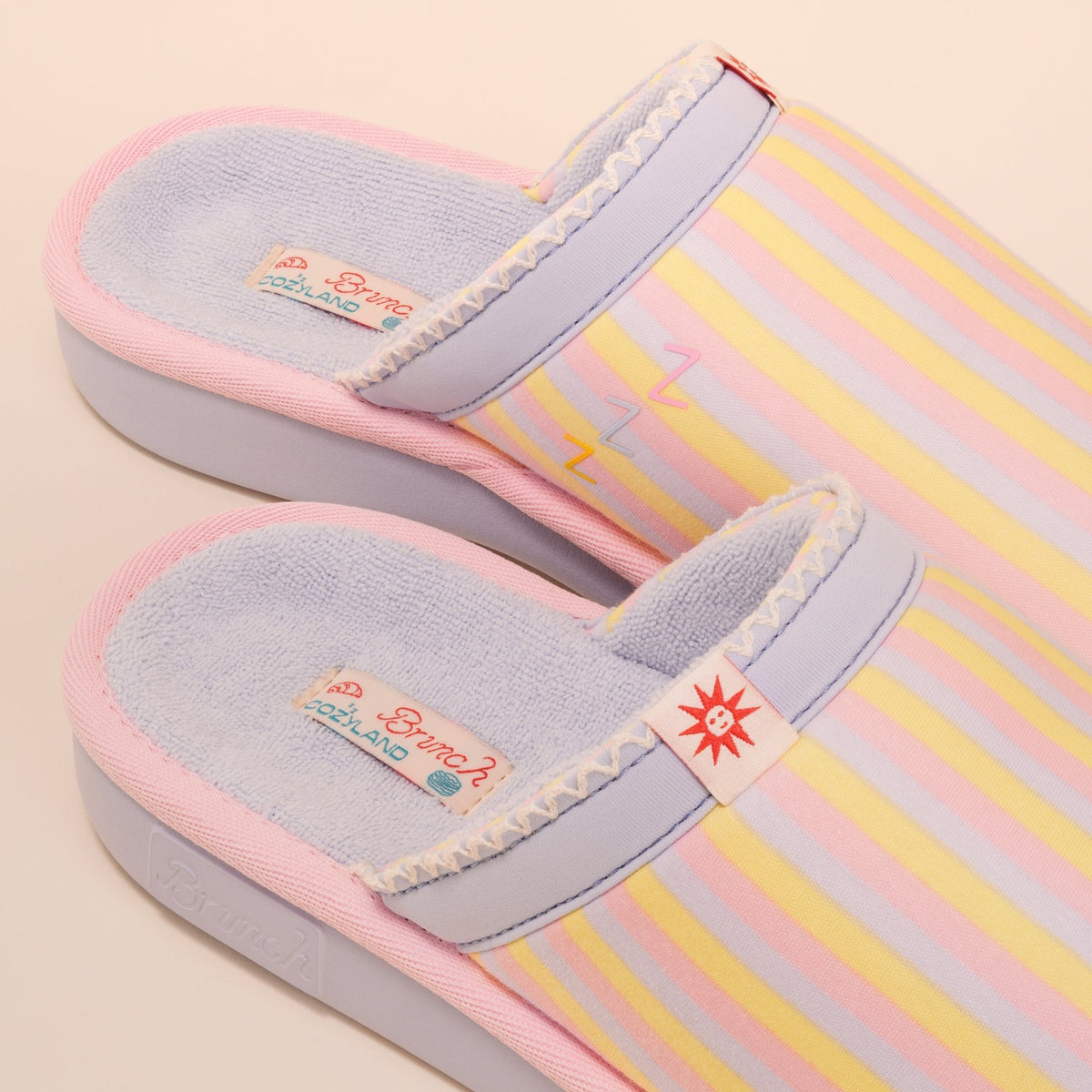 Top-down view of Cozyland Essential slipper, pastel rainbow stripes, soft terry lining, cushioned sole, sun motif tag on cream background