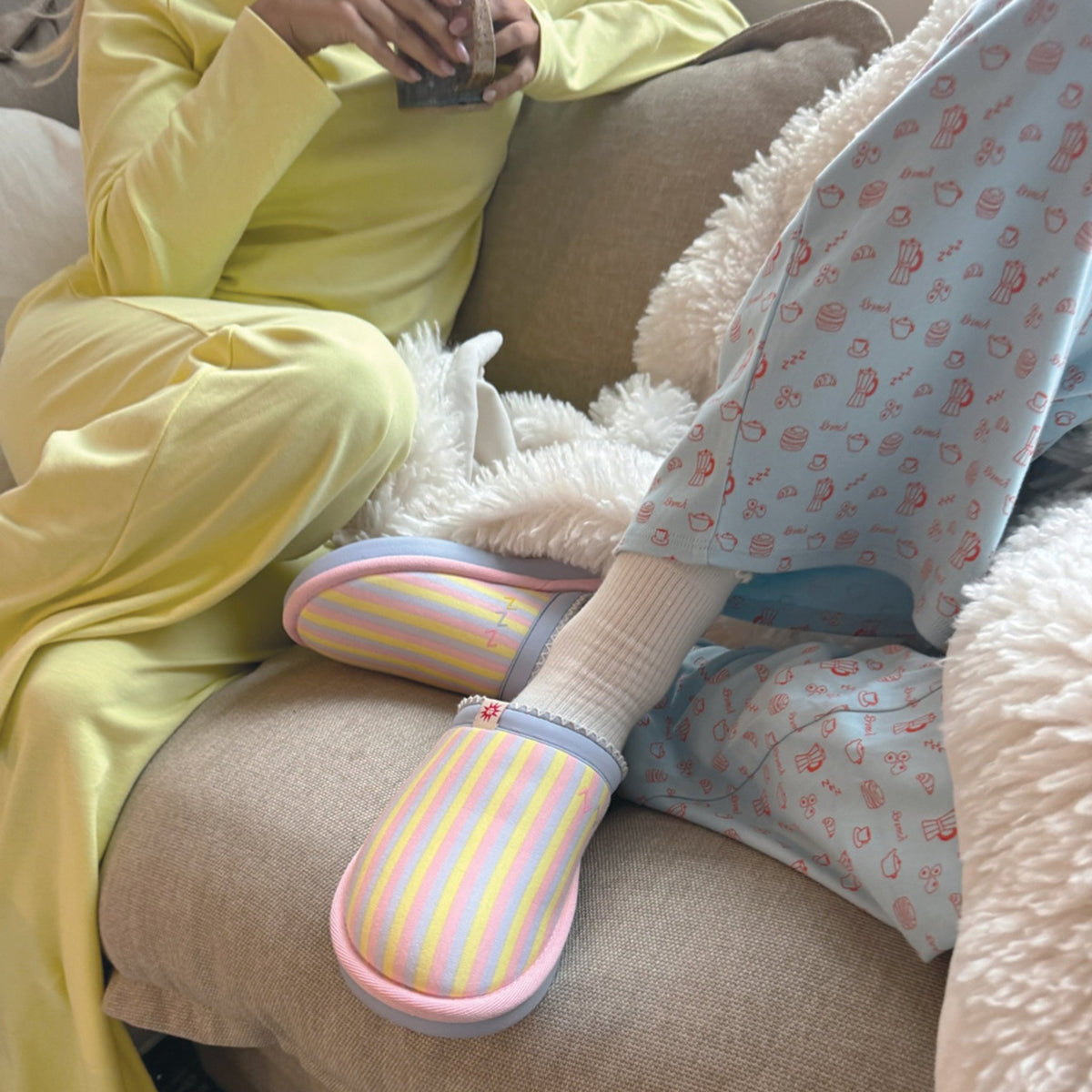 Three-quarter view of Cozyland Essential slipper showcasing pastel rainbow stripes and pink trim on a beige sofa
