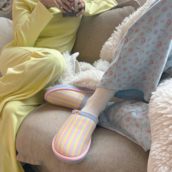 Three-quarter view of Cozyland Essential slipper showcasing pastel rainbow stripes and pink trim on a beige sofa