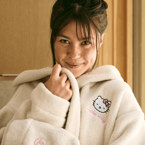Front view of cream Essential Robe on model showing embroidered Hello Kitty chest, hood, oversized fit, and terry texture