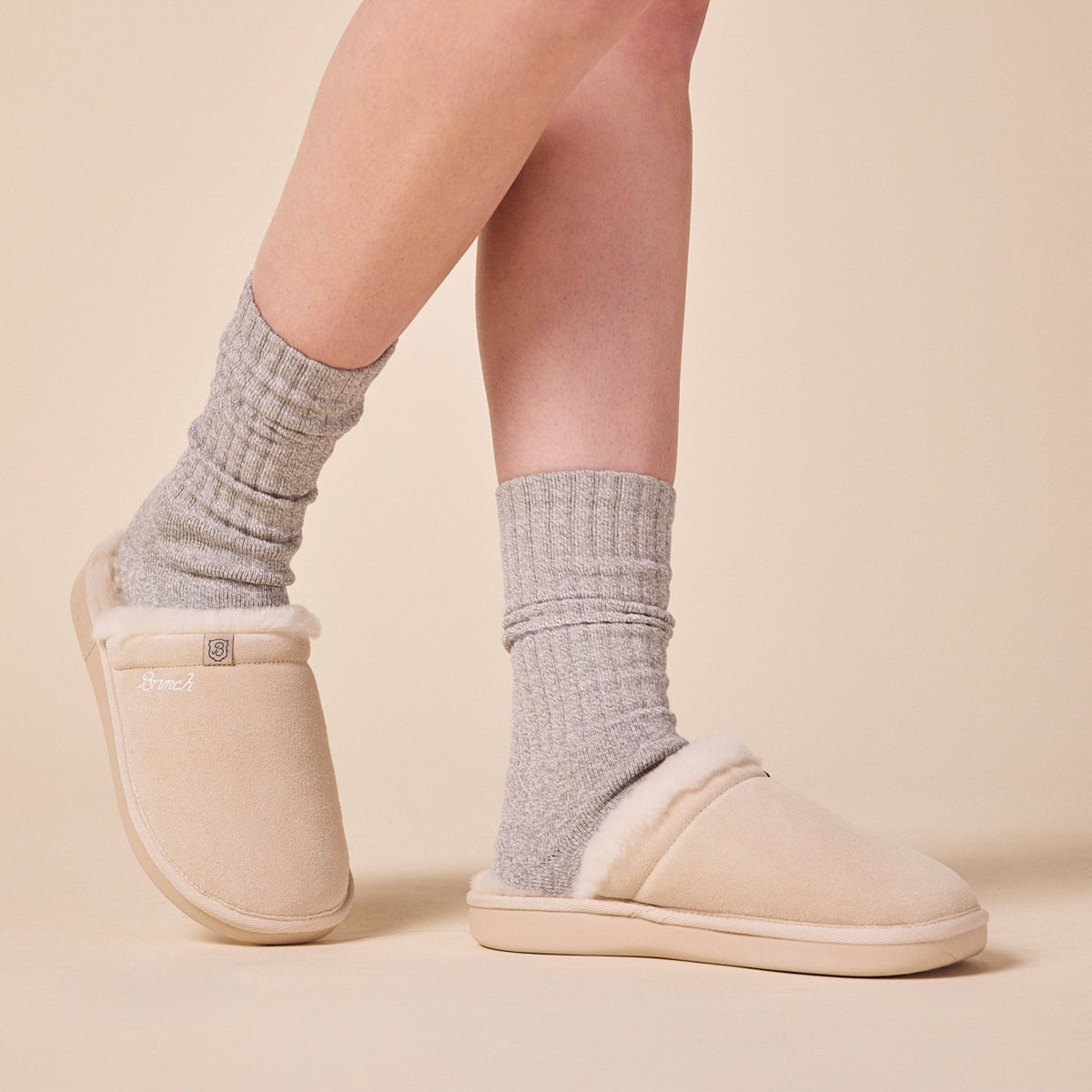 Side view of Essential Shearling suede slipper on model showing cream-beige suede upper, plush shearling lining, low-back slip-on