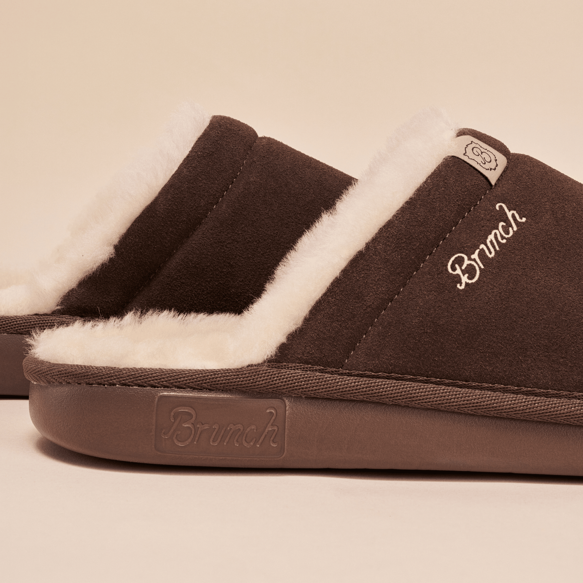 Close up side view of brown suede Essential Shearling slipper showing plush white shearling lining, Brunch embroidered logo and molded sole on beige background