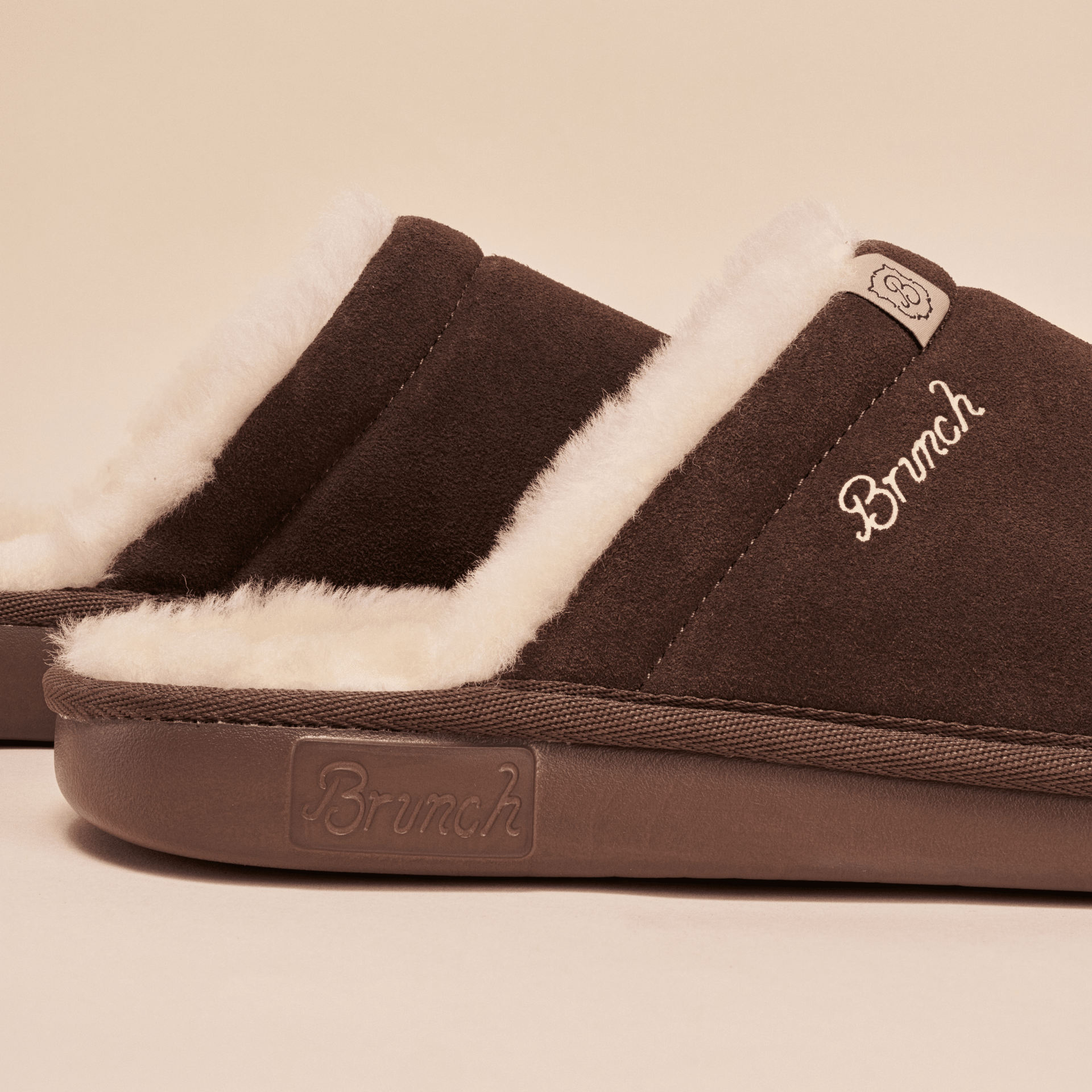 Close up side view of brown suede Essential Shearling slipper showing plush white shearling lining, Brunch embroidered logo and molded sole on beige background