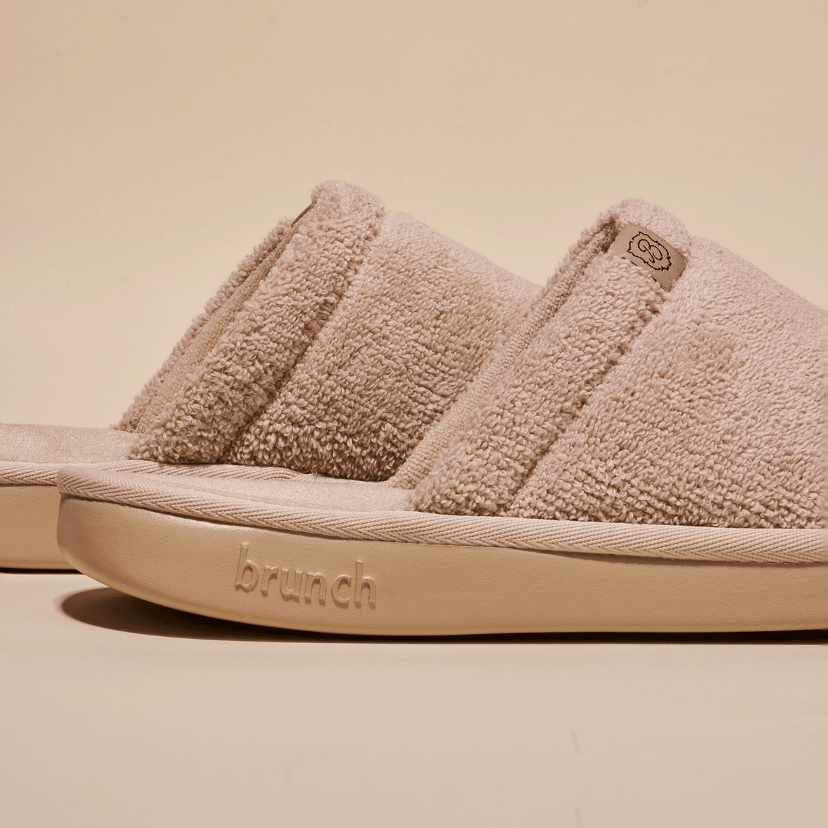 Side view of beige Essential Slipper showing soft terry upper, open-toe slide silhouette and embossed Brunch sole on neutral background