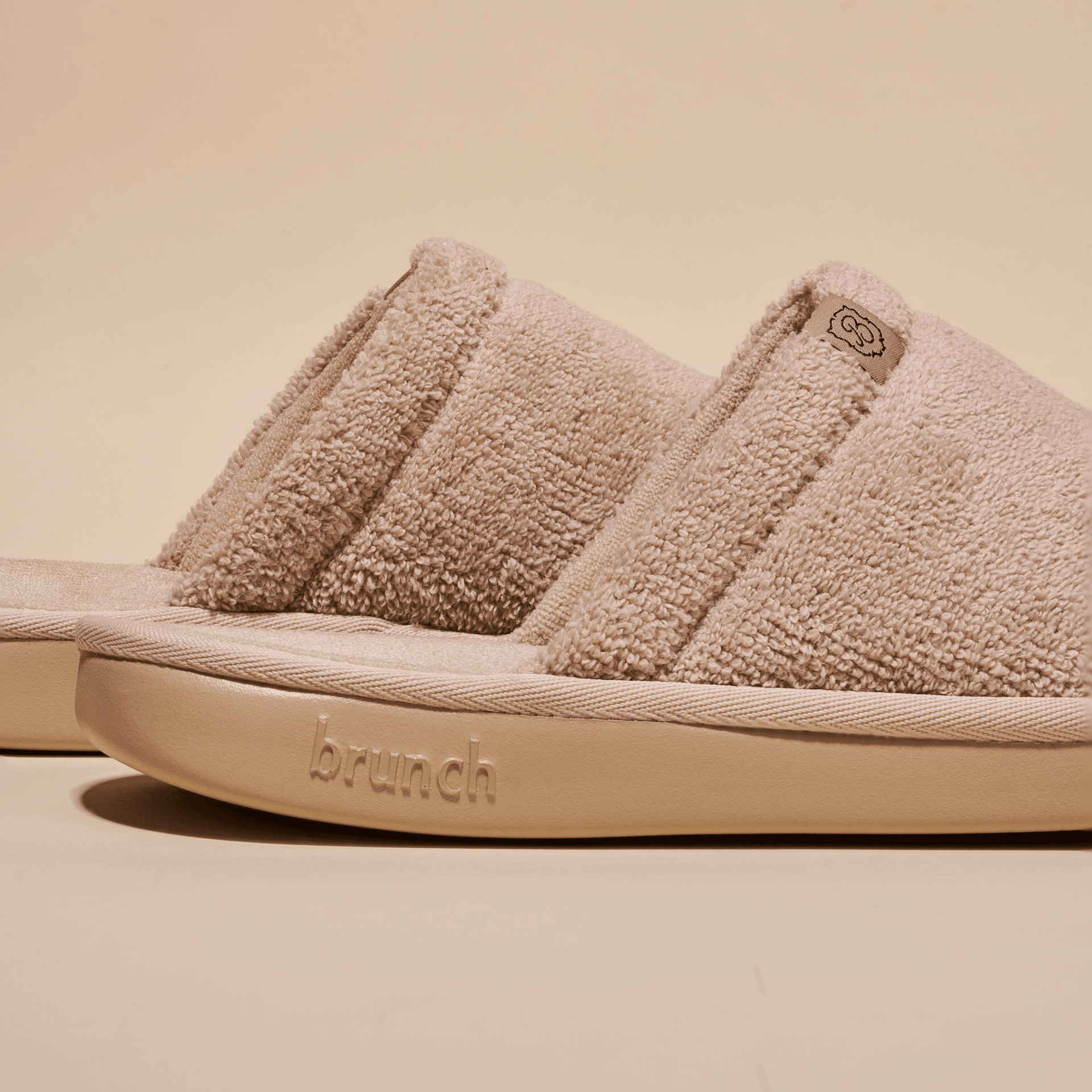 Side view of beige Essential Slipper showing soft terry upper, open-toe slide silhouette and embossed Brunch sole on neutral background