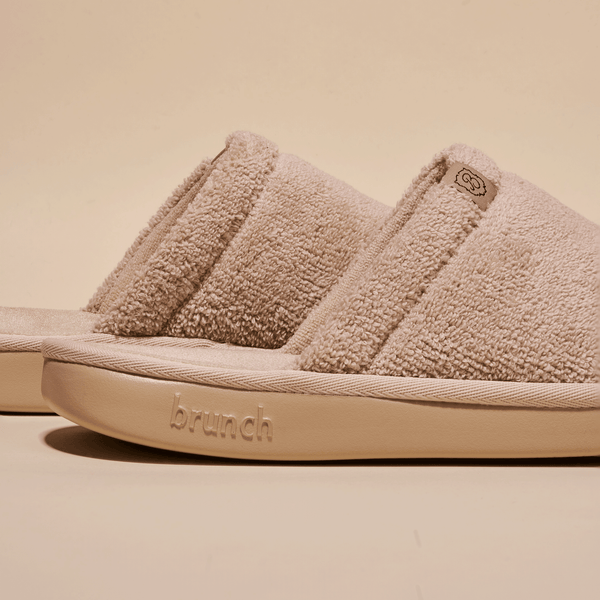 Side view of beige Essential Slipper showing soft terry upper, open-toe slide silhouette and embossed Brunch sole on neutral background