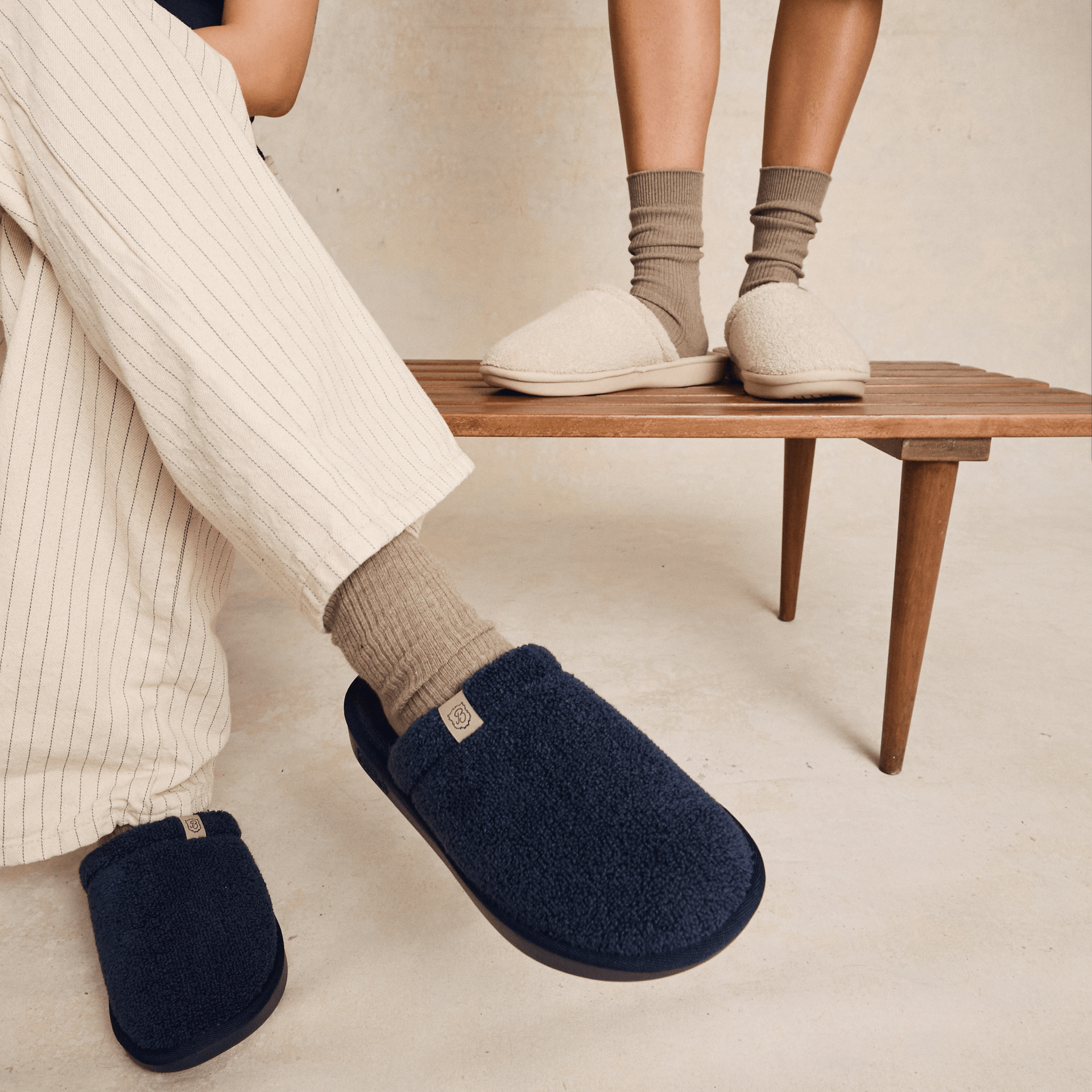Three-quarter view of Essential slipper in navy and beige showing plush sherpa upper and slip-on back on wooden bench