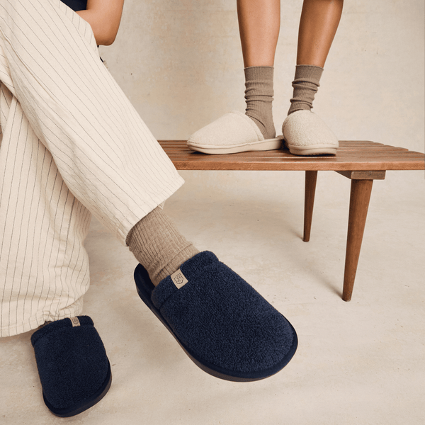 Three-quarter view of Essential slipper in navy and beige showing plush sherpa upper and slip-on back on wooden bench