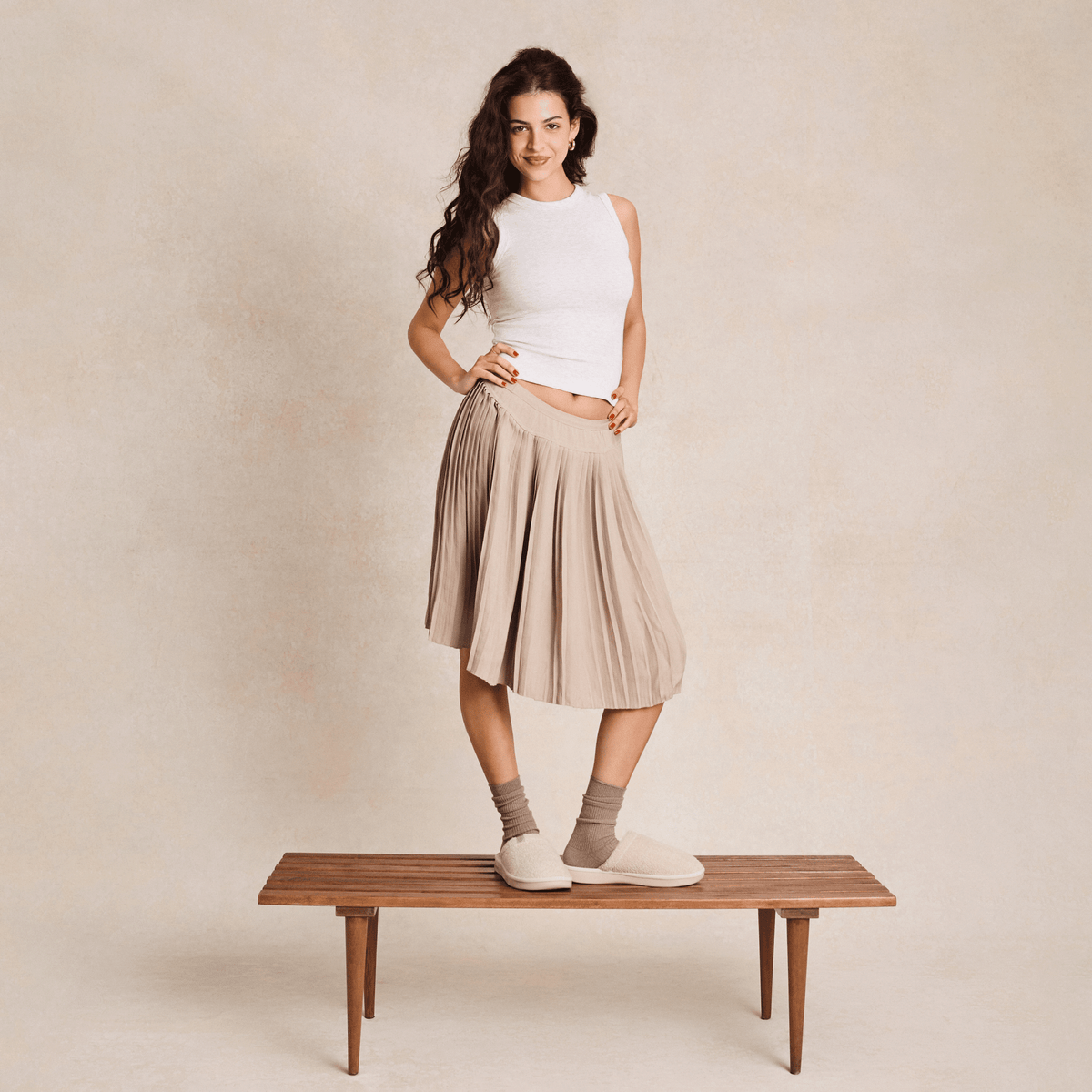 Woman standing on a bench wearing cream slippers and beige socks, pleated tan skirt and white sleeveless top against a neutral backdrop