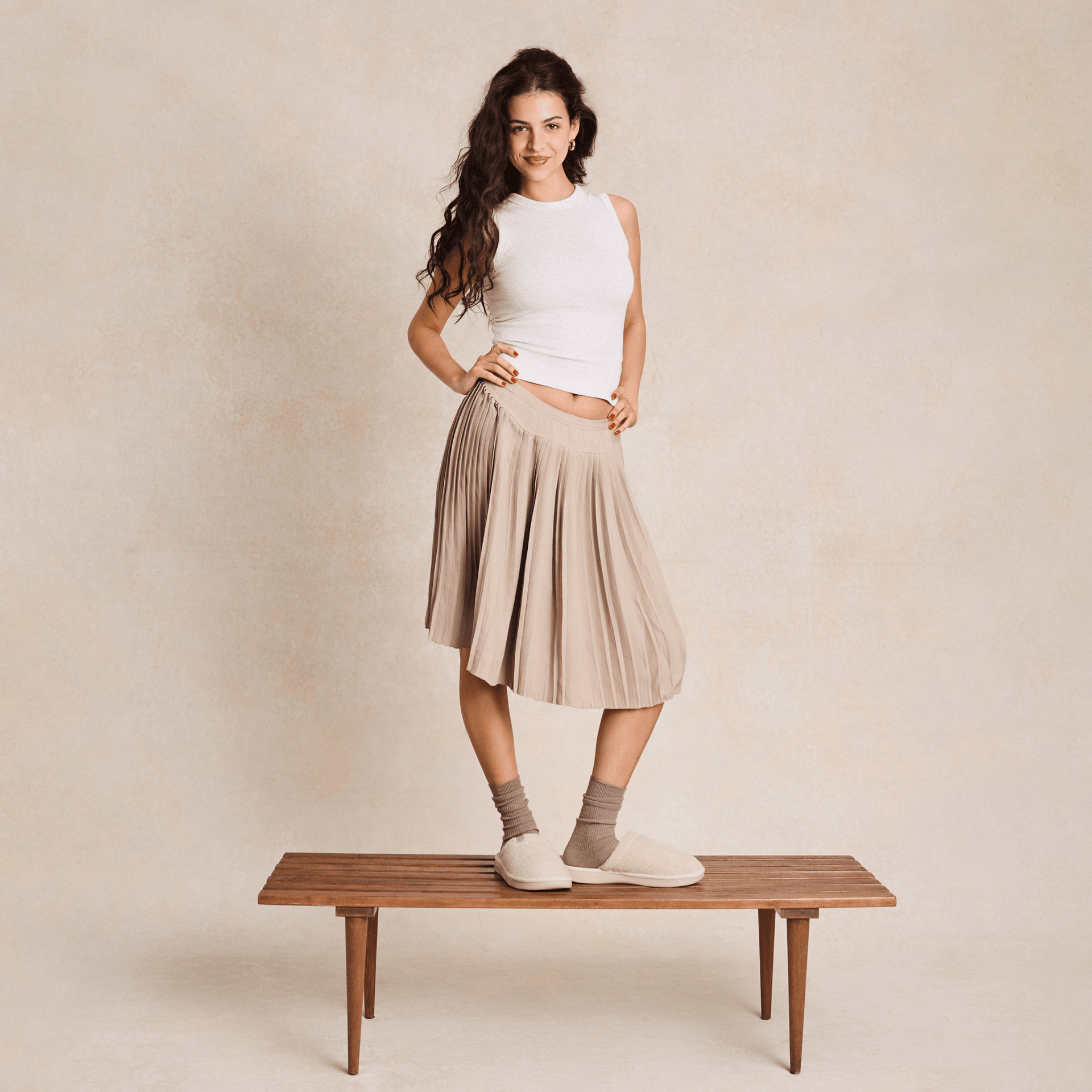 Woman standing on a bench wearing cream slippers and beige socks, pleated tan skirt and white sleeveless top against a neutral backdrop