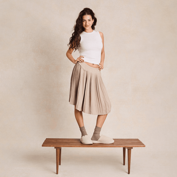 Woman standing on a bench wearing cream slippers and beige socks, pleated tan skirt and white sleeveless top against a neutral backdrop