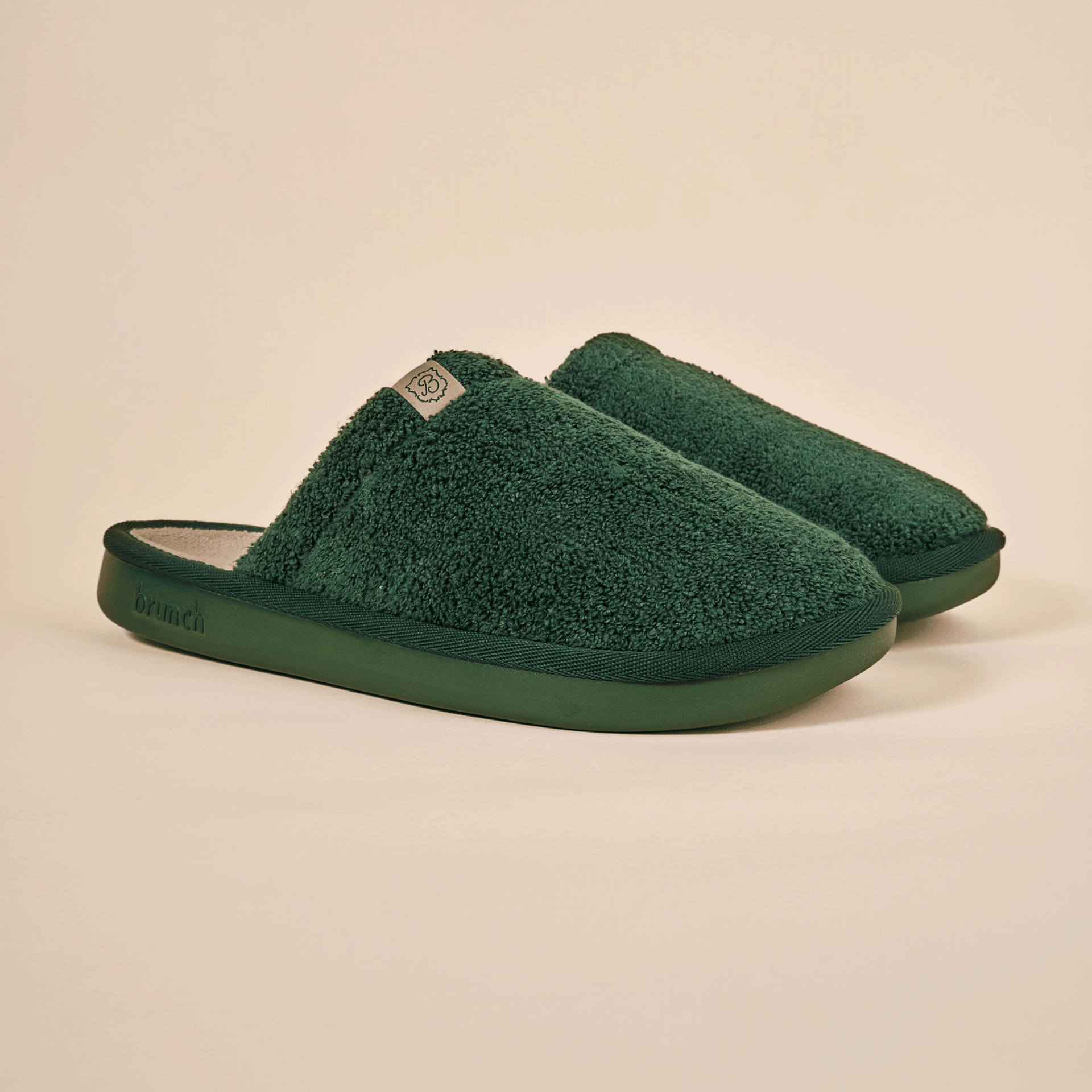 Three-quarter view of Essential Slipper in dark green terry fabric showing plush upper and molded green sole on cream background