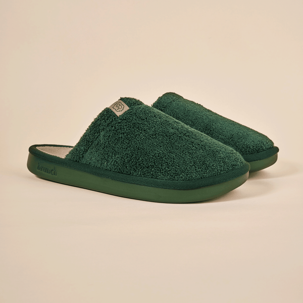 Three-quarter view of Essential Slipper in dark green terry fabric showing plush upper and molded green sole on cream background
