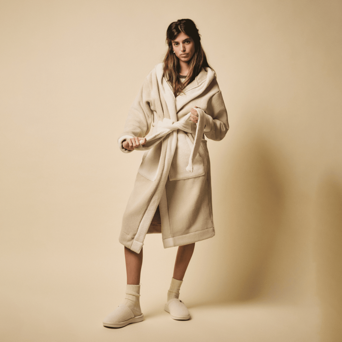 Three-quarter view of cream Essential Slipper on model showing plush fleece upper and low rubber sole on beige backdrop