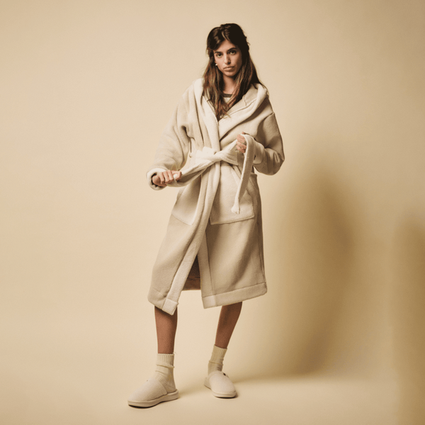Three-quarter view of cream Essential Slipper on model showing plush fleece upper and low rubber sole on beige backdrop