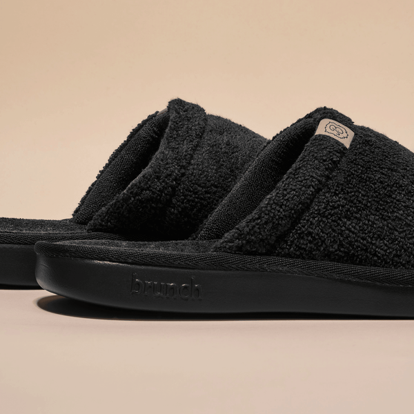 Close-up side view of Essential slipper showcasing black terry-cloth upper, open-toe slide design, and embossed brunch logo on the sole with beige background