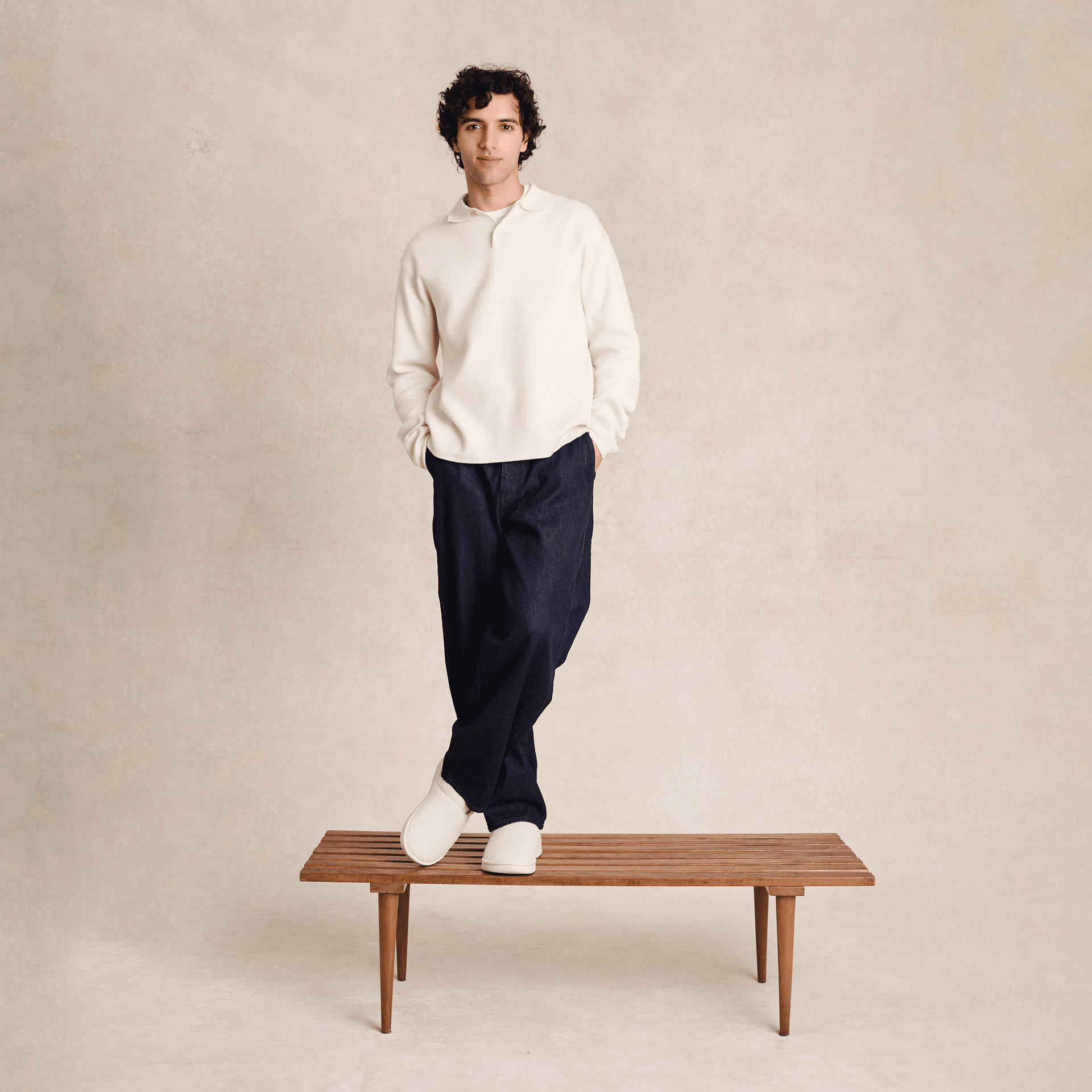 Model standing on wooden bench wearing cream Brunch Essential slipper and relaxed dark pants against neutral studio backdrop