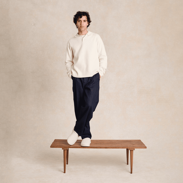 Model standing on wooden bench wearing cream Brunch Essential slipper and relaxed dark pants against neutral studio backdrop