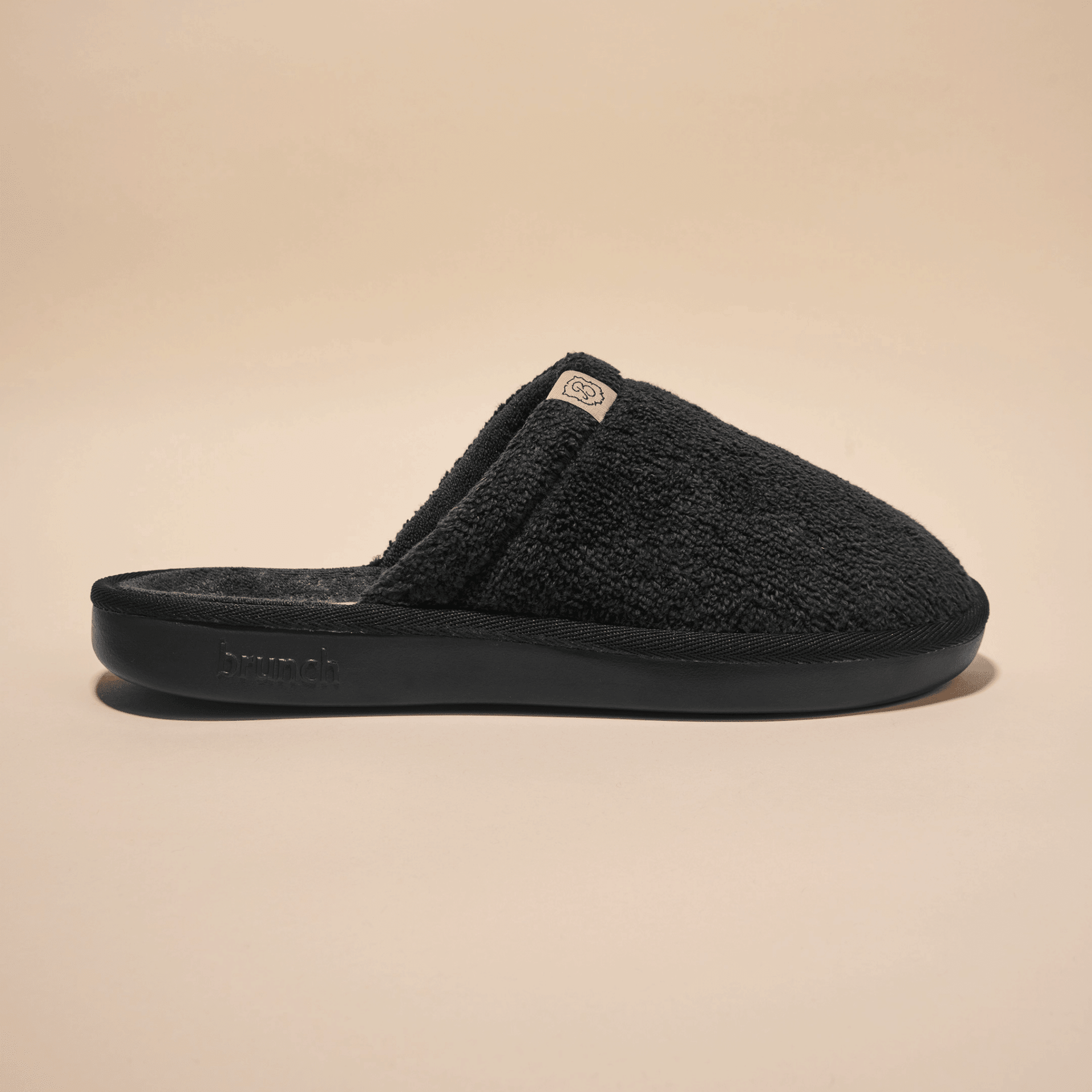 Side view of Essential Slipper, black, showing terry-cloth upper, open-back slip-on design and molded sole on beige background