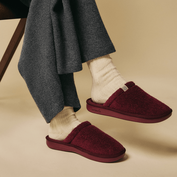 Side view of burgundy Essential Slipper with plush sherpa upper, backless slip on and branded sole on beige background