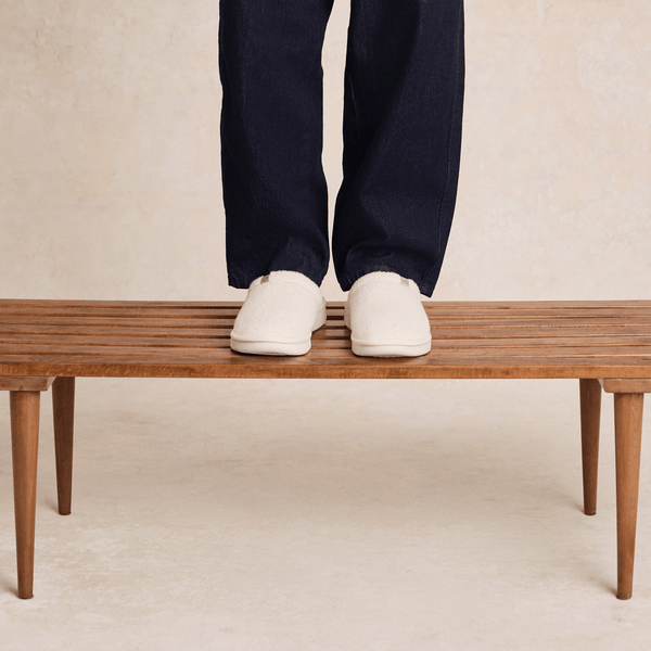 Front view of Essential Slipper in cream sherpa showcasing low-profile slip-on silhouette and cushioned sole on a wooden bench