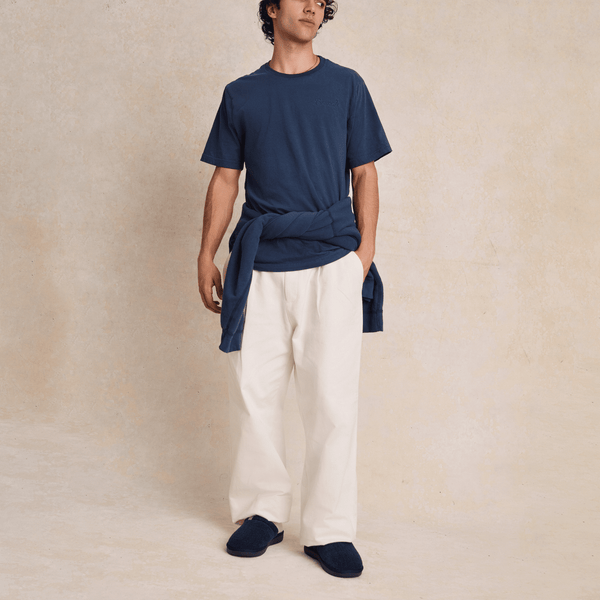 Man standing with hands in pockets wearing navy slippers, navy tee and white wide-leg pants on neutral studio background