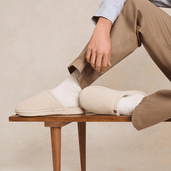 Side view of Essential slipper in cream showing plush boucle upper and low-profile sole, worn with white socks on wooden bench