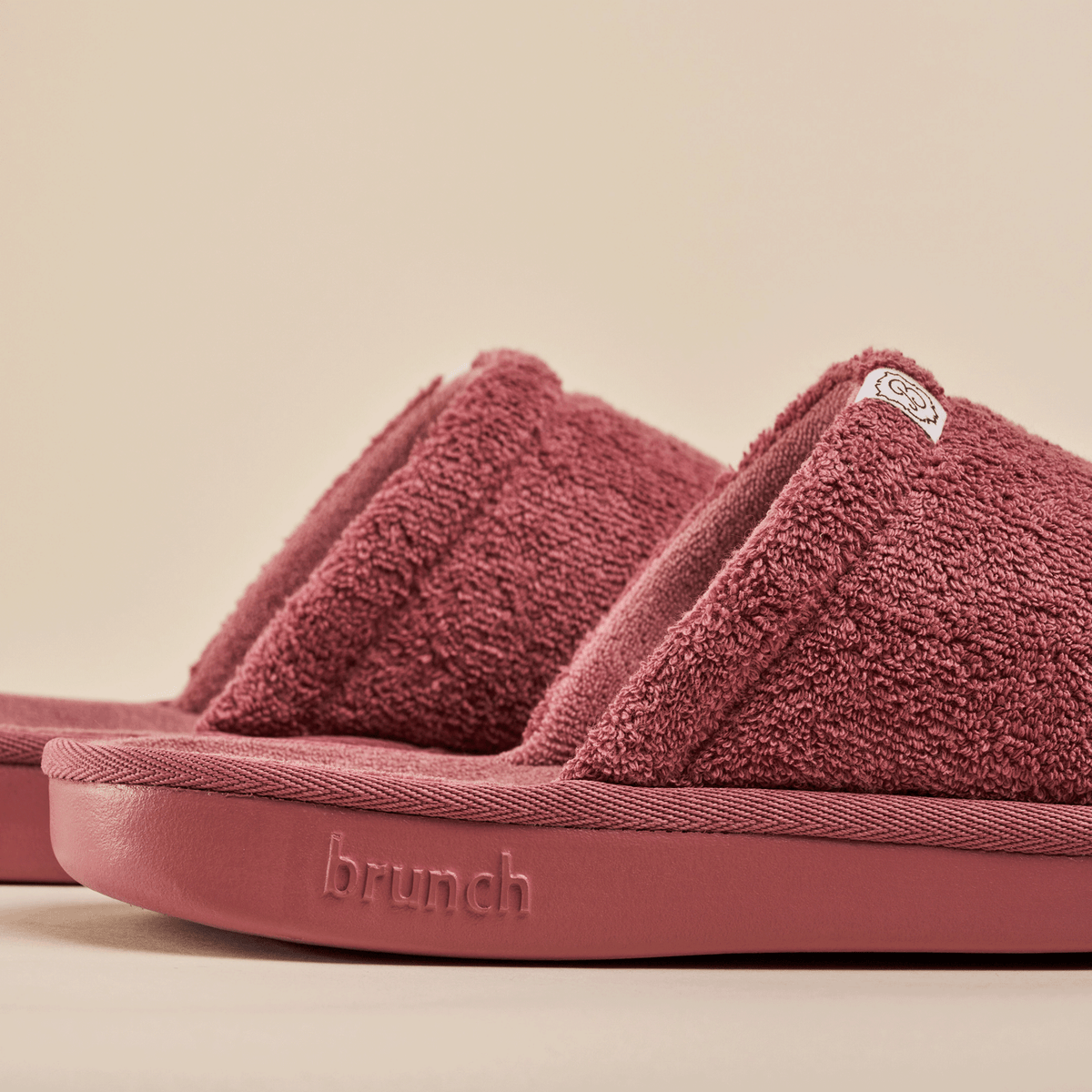 Close-up of Essential slipper in rose showing terry-cloth upper, open-toe design and embossed brunch logo on beige background