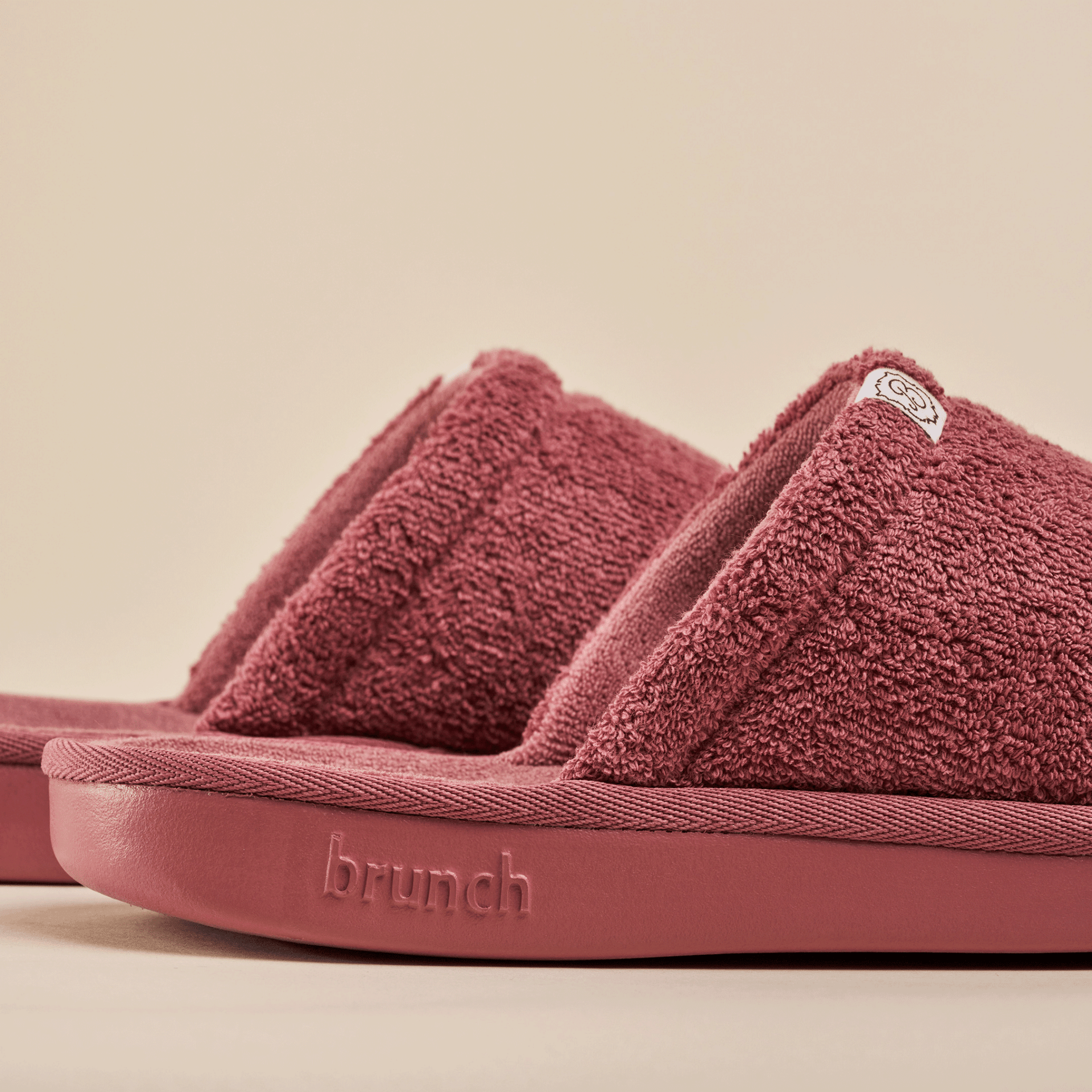 Close-up of Essential slipper in rose showing terry-cloth upper, open-toe design and embossed brunch logo on beige background