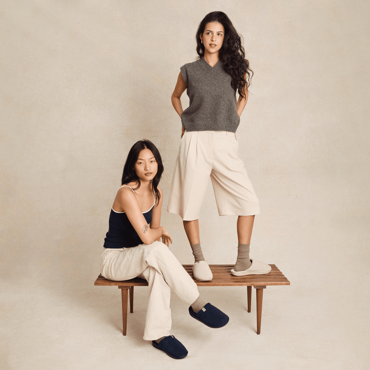 Two women modeling Brunch Essential slippers in navy and beige on a wooden bench against a neutral studio backdrop