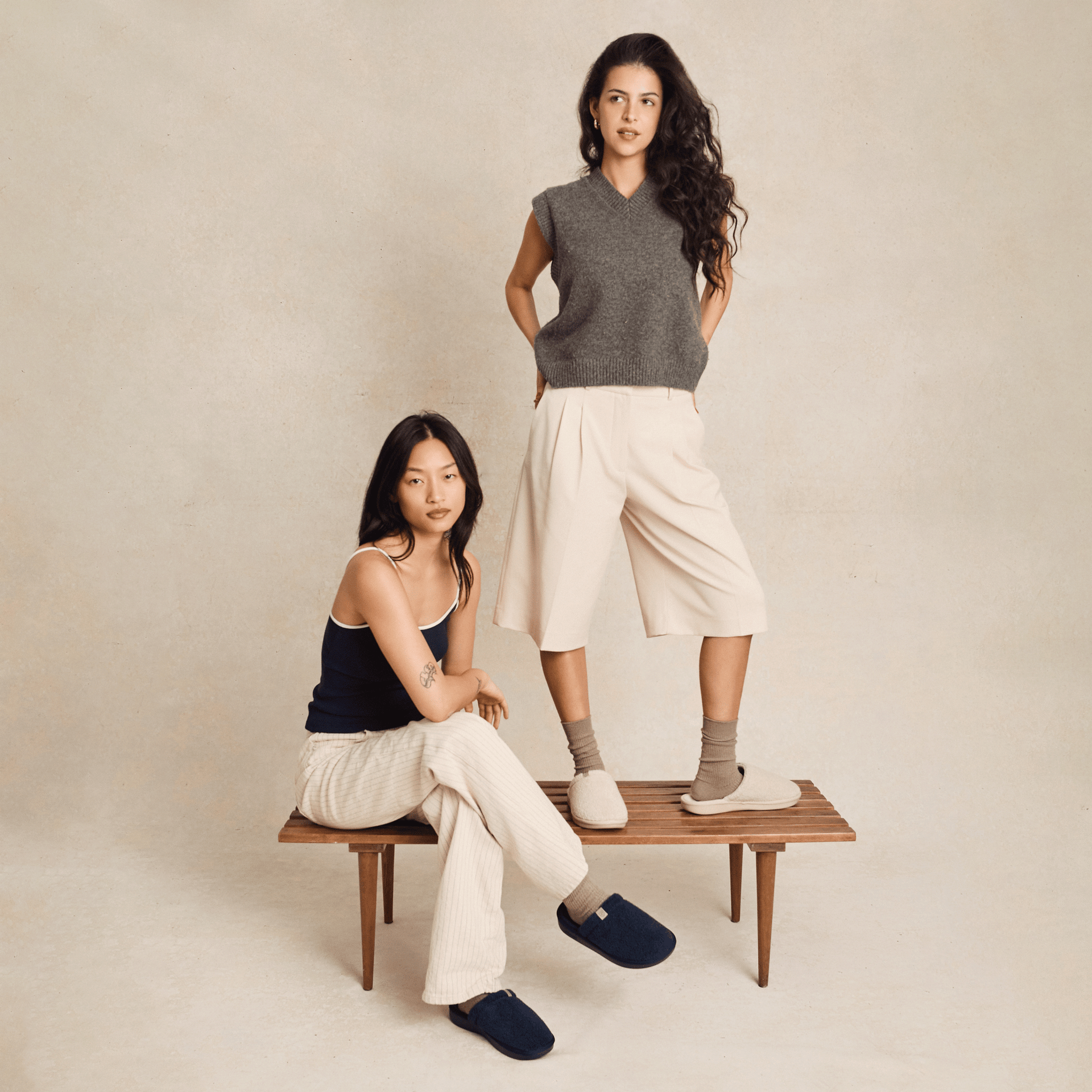 Two women modeling Brunch Essential slippers in navy and beige on a wooden bench against a neutral studio backdrop