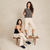 Two women modeling Brunch Essential slippers in navy and beige on a wooden bench against a neutral studio backdrop
