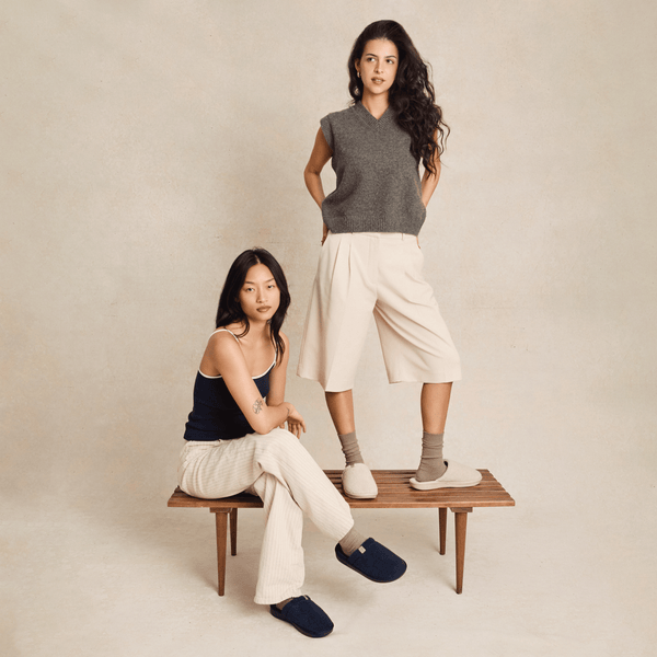 Two women modeling Brunch Essential slippers in navy and beige on a wooden bench against a neutral studio backdrop