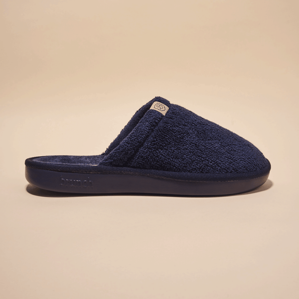 Side view of navy Essential slipper in terry cloth showing closed toe and cushioned molded sole embossed with Brunch logo on beige background